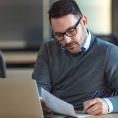 Mann sitzt am Tisch vor einem Notebook und schreibt etwas auf