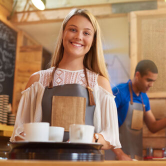 Eine junge Mitarbeiterin in einem Cafe lächelt in die Kamera, als sie sich darauf vorbereitet, eine Bestellung an einen Tisch zu bringen.