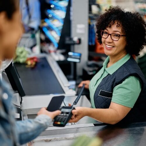 Glückliche Kassiererin hält Kreditkartenlesegerät in der Hand, während der Kunde im Supermarkt mit dem Smartphone bezahlt.
