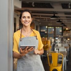 Eine fröhliche Kellnerin, die am Eingang eines Cafés steht und ein Tablet in der Hand hält.