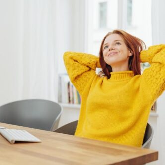 Porträt einer jungen, verträumten und entspannten Frau im gelben Pullover, die vor dem Computer sitzt.
