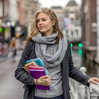 Junge Frau mit Schal und Mantel, die Notizbücher und Hefte trägt, steht auf einer Brücke in einer urbanen Umgebung.