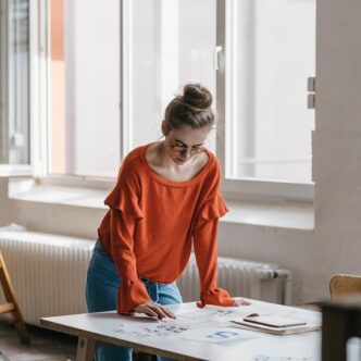 Junge Frau mit hochgesteckten Haaren und rotem Pullover steht an einem Tisch und arbeitet an kreativen Entwürfen.