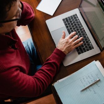 Ein Mann in einem roten Pullover sitzt an einem Schreibtisch und arbeitet an einem Laptop. Neben dem Laptop liegen ein Notizblock und ein Stift. Der Mann schaut auf den Bildschirm und tippt auf der Tastatur.