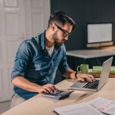 Mann mit Brille arbeitet am Laptop und nutzt einen Taschenrechner, auf dem Schreibtisch liegen Unterlagen und eine Kaffeetasse.
