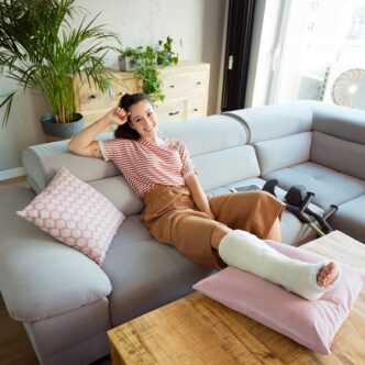 Junge Frau mit Gipsbein sitzt lächelnd auf einem Sofa und hat das verletzte Bein auf einem Kissen hochgelegt. Neben ihr liegen Krücken.