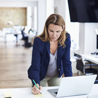 Frau in Businesskleidung steht an einem Schreibtisch, arbeitet am Laptop und macht sich Notizen.