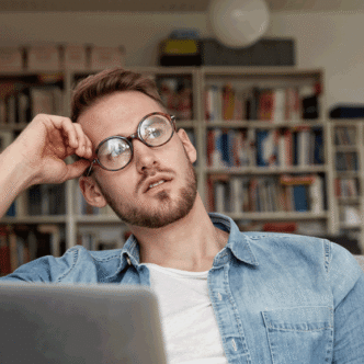 Ein junger Mann mit Brille sitzt gedankenvertieft am Laptop im Arbeitszimmer.