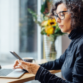Frau mit Brille arbeitet am Laptop und schaut auf ihr Smartphone, im Hintergrund ein Blumenstrauß.
