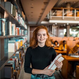 Eine junge Frau steht in einer Bibliothek und hält Bücher in der Hand
